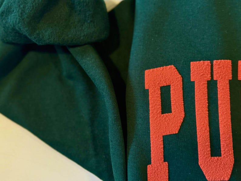 May include: Close-up of a dark green sweatshirt with the word "PUT" in a bold, orange, textured font. The fabric is folded, showing the texture and color variations of the material.