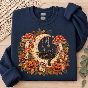 May include: A navy blue crewneck sweatshirt featuring a graphic print of a black cat curled up on a crescent moon, surrounded by red and white spotted mushrooms, two jack-o'-lantern pumpkins, autumn leaves, and hanging star ornaments.
