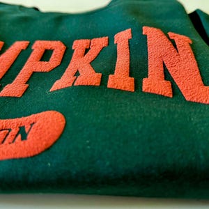 May include: A dark green sweatshirt with the word "PUMPKIN" in orange, textured lettering across the chest. Below, an orange patch reads "SON". The fabric appears soft and slightly worn.