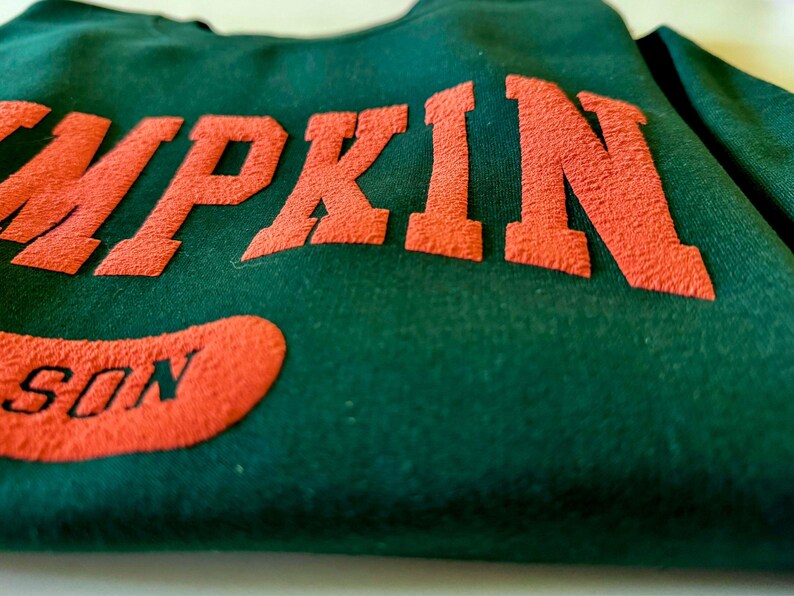 May include: A dark green sweatshirt with the word "PUMPKIN" in orange, textured lettering across the chest. Below, an orange patch reads "SON". The fabric appears soft and slightly worn.