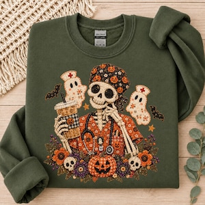 May include: An olive green crewneck sweatshirt featuring a graphic of a skeleton dressed as a nurse with a stethoscope, holding a coffee cup. The design includes two ghost figures, bats, a jack-o'-lantern, and floral accents in orange and purple.