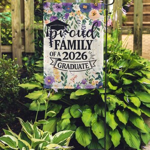 May include: A garden flag with the text "Proud Family of a 2026 Graduate" in black script. The flag features a graduation cap and is decorated with a floral border of purple, blue, and peach flowers. The flag is displayed on a black metal stand.
