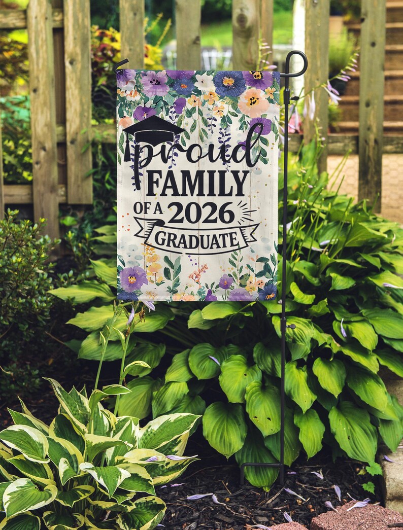 May include: A garden flag with the text "Proud Family of a 2026 Graduate" in black script. The flag features a graduation cap and is decorated with a floral border of purple, blue, and peach flowers. The flag is displayed on a black metal stand.