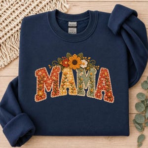 Puede incluir: Una sudadera azul marino de cuello redondo con la palabra MAMA en grandes letras de tela aplicadas. Cada letra está rellena con diferentes patrones florales y de cuadros en naranja, amarillo y verde, rematada con flores de tela en relieve.