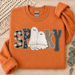 Peut inclure: Un sweat-shirt orange à col rond avec le mot SPOOKY appliqué sur la poitrine. Les lettres sont confectionnées dans divers tissus, notamment du vichy noir et blanc, un imprimé floral noir et deux fantômes en dentelle blanche.