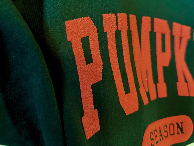 May include: Close-up of a dark green sweatshirt with the word "PUMPKIN" in large, orange, textured letters. Below, the word "SEASON" is printed in a matching orange oval.