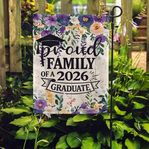 May include: A garden flag with a floral design and the text "Proud Family of a 2026 Graduate." The flag features a graduation cap and a banner. The flowers are in shades of purple, yellow, and orange.