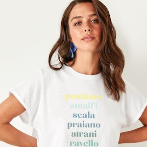 Puede incluir: Camiseta blanca con un estampado gr&aacute;fico colorido de los nombres de ciudades italianas: Positano, Amalfi, Scala, Praiano, Atrani, Ravello.