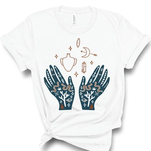 Puede incluir: Camiseta blanca con una ilustraci&oacute;n azul y marr&oacute;n de dos manos con s&iacute;mbolos m&iacute;sticos, incluyendo una luna creciente, un cristal, un jarr&oacute;n y serpientes.
