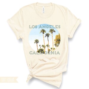 May include: A soft cream t-shirt featuring a circular graphic of palm trees against a blue sky. The text "Los Angeles California" is printed in green around the graphic.