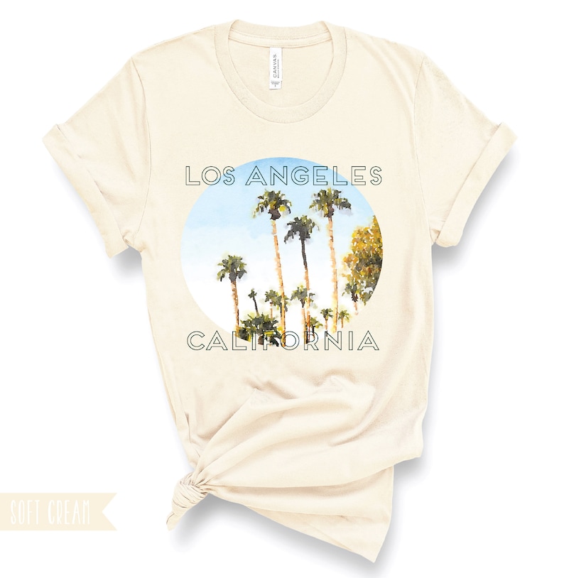 May include: A soft cream t-shirt featuring a circular graphic of palm trees against a blue sky. The text "Los Angeles California" is printed in green around the graphic.