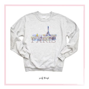 May include: A gray sweatshirt with a watercolor illustration of the Eiffel Tower and other Parisian landmarks. The word "PARIS" is printed in pink below the illustration.