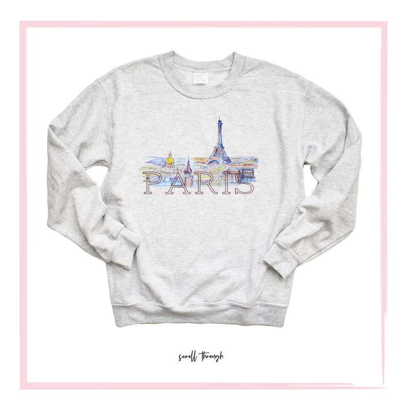 May include: A gray sweatshirt with a watercolor illustration of the Eiffel Tower and other Parisian landmarks. The word "PARIS" is printed in pink below the illustration.