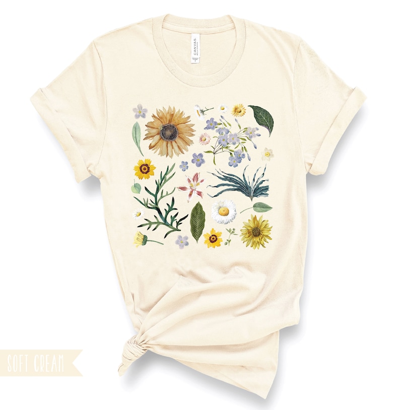 May include: A cream colored t-shirt with a floral print featuring sunflowers, daisies, and other flowers in shades of yellow, blue, and green.