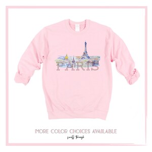 May include: A pink sweatshirt with a watercolor illustration of the Eiffel Tower and other Parisian landmarks. The word "PARIS" is printed in black below the illustration. The text "MORE COLOR CHOICES AVAILABLE scroll through" is printed below the image.