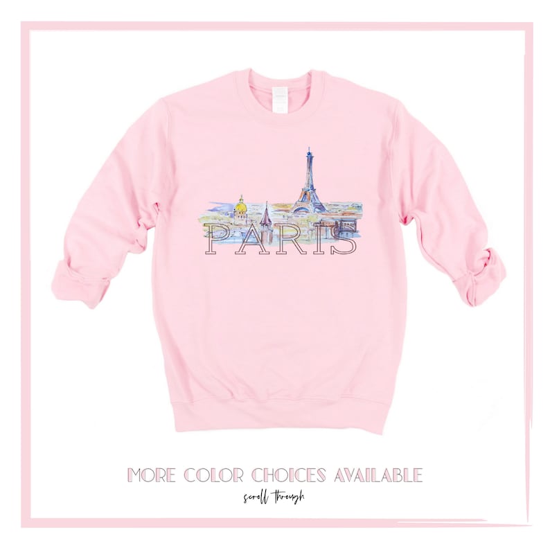 May include: A pink sweatshirt with a watercolor illustration of the Eiffel Tower and other Parisian landmarks. The word "PARIS" is printed in black below the illustration. The text "MORE COLOR CHOICES AVAILABLE scroll through" is printed below the image.
