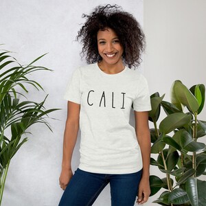 Pu&ograve; includere: Una donna indossa una maglietta grigia chiaro con la scritta "CALI" stampata in nero sul davanti. &Egrave; in piedi davanti a un muro bianco con piante su entrambi i lati.