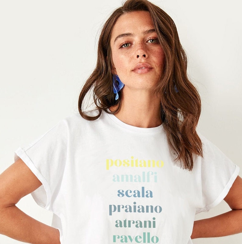 Puede incluir: Camiseta blanca con un estampado gr&aacute;fico colorido de los nombres de ciudades italianas: Positano, Amalfi, Scala, Praiano, Atrani, Ravello.