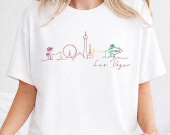 May include: White t-shirt with a vibrant line drawing of the Las Vegas skyline. The design includes the Eiffel Tower, a Ferris wheel, and the "Welcome to Las Vegas" sign. The words "Las Vegas" are written below the skyline.