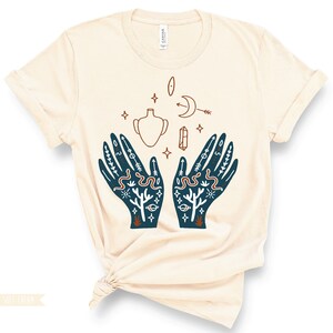 Puede incluir: Una camiseta beige con una ilustraci&oacute;n azul de dos manos con varios s&iacute;mbolos, incluyendo una luna creciente, una estrella, un cristal, un jarr&oacute;n y una serpiente. Los s&iacute;mbolos est&aacute;n en un estilo m&iacute;stico.