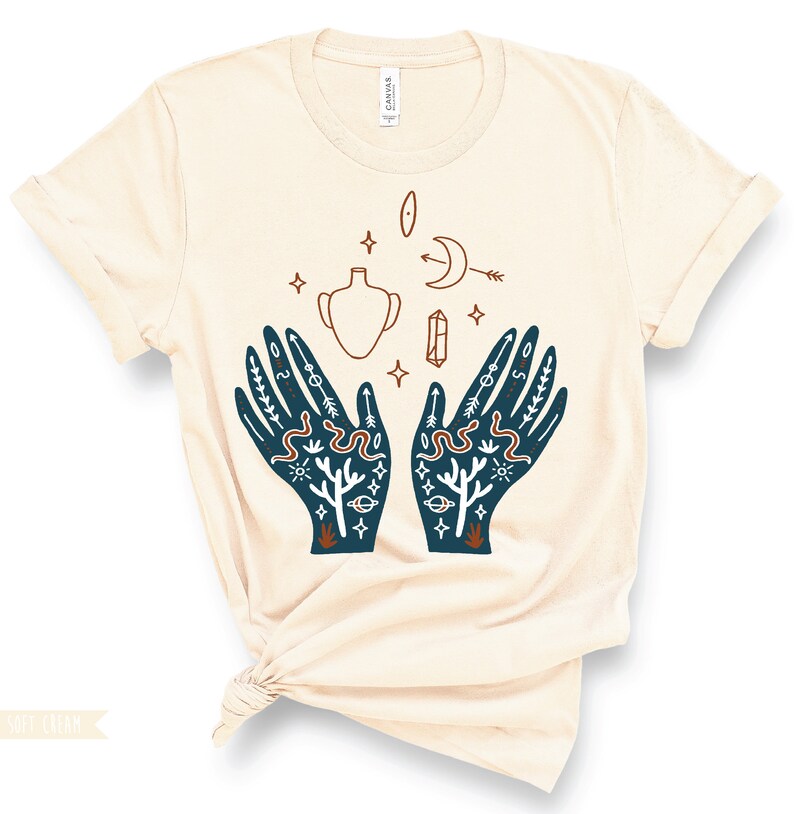 Puede incluir: Una camiseta beige con una ilustraci&oacute;n azul de dos manos con varios s&iacute;mbolos, incluyendo una luna creciente, una estrella, un cristal, un jarr&oacute;n y una serpiente. Los s&iacute;mbolos est&aacute;n en un estilo m&iacute;stico.