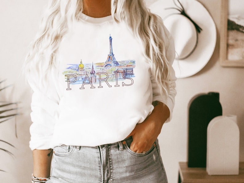 May include: White sweatshirt with a watercolor illustration of the Eiffel Tower and the word "PARIS" printed in pink.