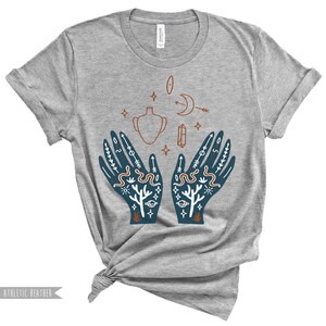 Puede incluir: Camiseta gris con un dise&ntilde;o de dos manos con varios s&iacute;mbolos y dise&ntilde;os, incluyendo una luna creciente, una estrella, una serpiente, un jarr&oacute;n, un cristal y un sol.