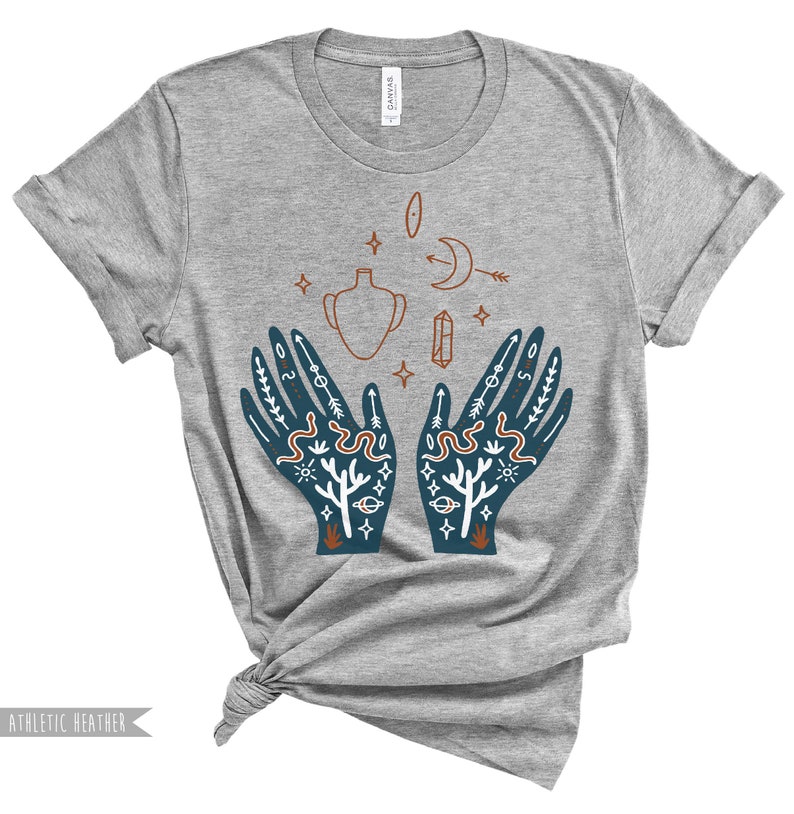 Puede incluir: Camiseta gris con un dise&ntilde;o de dos manos con varios s&iacute;mbolos y dise&ntilde;os, incluyendo una luna creciente, una estrella, una serpiente, un jarr&oacute;n, un cristal y un sol.