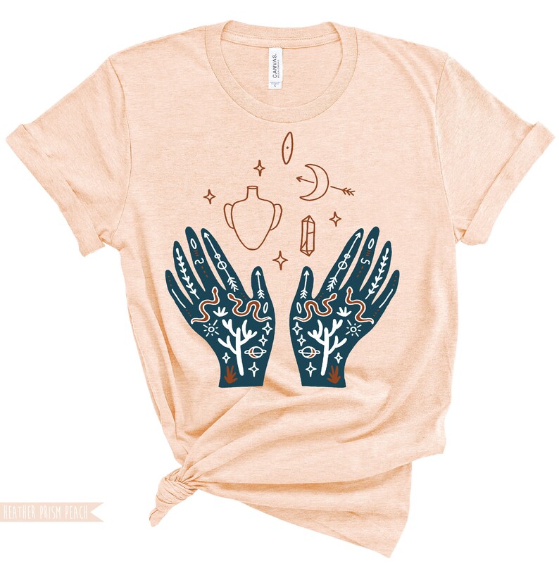 Puede incluir: Una camiseta de color melocot&oacute;n con un dise&ntilde;o gr&aacute;fico de dos manos con intrincados patrones, incluyendo serpientes, estrellas, lunas y un jarr&oacute;n. Las manos se enfrentan, con las palmas hacia arriba, como si sostuvieran algo.
