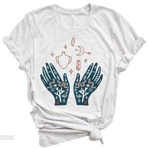 Puede incluir: Una camiseta gris claro con un dise&ntilde;o azul y naranja de dos manos con varios s&iacute;mbolos, incluyendo una luna creciente, un cristal, un jarr&oacute;n y serpientes.