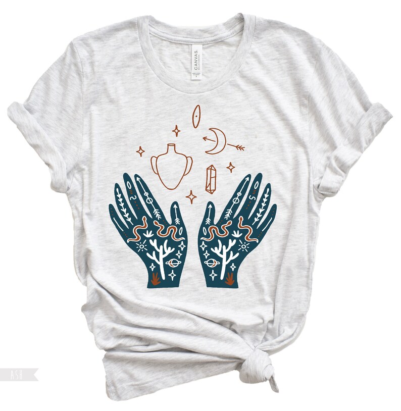 Puede incluir: Una camiseta gris claro con un dise&ntilde;o azul y naranja de dos manos con varios s&iacute;mbolos, incluyendo una luna creciente, un cristal, un jarr&oacute;n y serpientes.