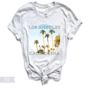May include: A heather grey t-shirt with a circular graphic of palm trees against a blue sky. The graphic says "Los Angeles California" in a teal font.