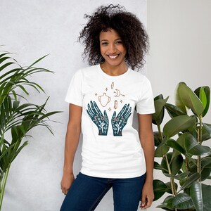 Puede incluir: Camiseta blanca con una ilustraci&oacute;n azul de dos manos sosteniendo una olla estilizada con una luna creciente, estrellas y otros s&iacute;mbolos.