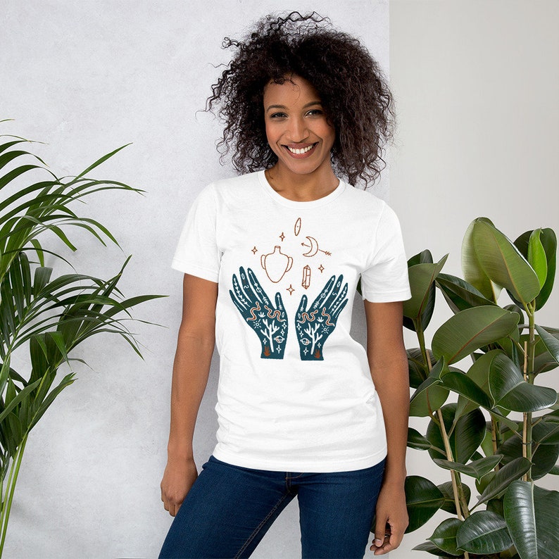 Puede incluir: Camiseta blanca con una ilustraci&oacute;n azul de dos manos sosteniendo una olla estilizada con una luna creciente, estrellas y otros s&iacute;mbolos.