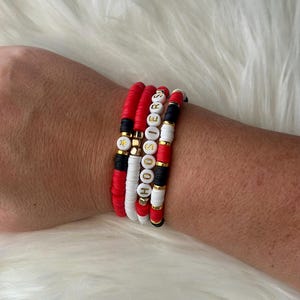 Puede incluir: Cuatro pulseras apiladas con cuentas rojas, blancas y negras. Las cuentas deletrean "HOOSIERS" en letras doradas.