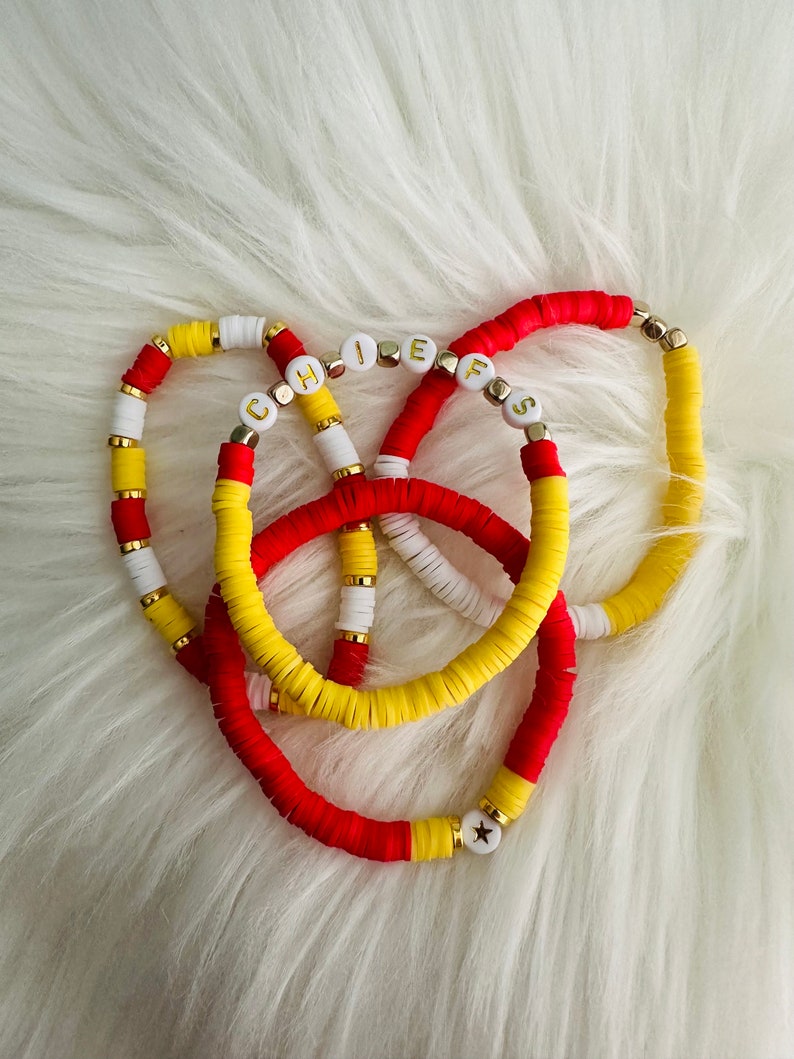 May include: Three colorful bracelets with white and yellow beads, and red and yellow beads. The bracelets have gold beads with white letters spelling out "CHIEFS" on them. One bracelet has a gold star bead.