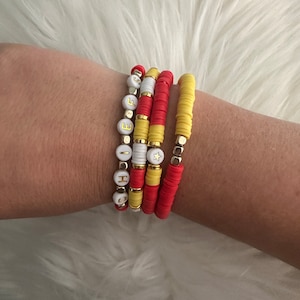 May include: A set of four bracelets with red and yellow beads. The bracelets are stacked on a person's wrist. One bracelet has white beads with gold letters spelling "HOPE" and a gold star charm.