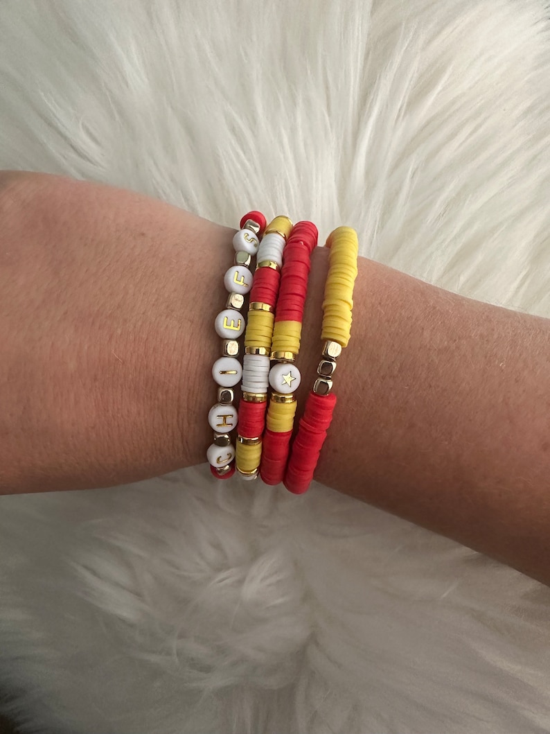 May include: A set of four bracelets with red and yellow beads. The bracelets are stacked on a person's wrist. One bracelet has white beads with gold letters spelling "HOPE" and a gold star charm.