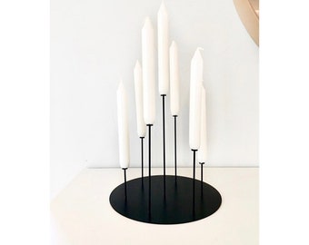 May include: A black metal candle holder with seven arms, each holding a white candle. The holder is on a white surface.