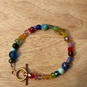 May include: A colorful beaded bracelet with a gold-colored toggle clasp. The bracelet features a variety of glass beads in shades of blue, green, yellow, red, and purple. The beads are different shapes and sizes, creating a unique and eye-catching accessory.