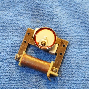 May include: A metallic electrical component featuring a rectangular frame with a copper-colored cylindrical coil at the base and a circular silver-toned motor housing with a small central gear positioned at the top center of the frame.