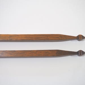 May include: Two wooden magnetic poster hangers. Each hanger is rectangular with a pointed end and a decorative finial. The wood has a warm brown tone and visible grain. The hangers are designed to display artwork or posters.