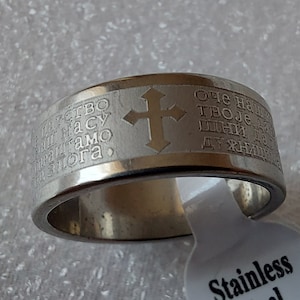 Pode incluir: Um anel de aço inoxidável prateado com uma cruz e texto gravado. O texto diz "Отец наш, Иисус Христос, Сын Божий, спаси нас".