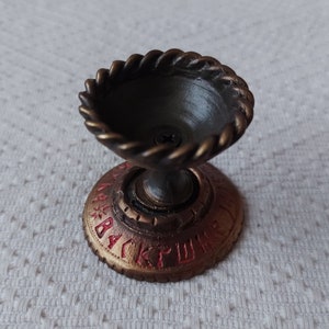 May include: A small, gold-coloured metal candle holder with a round base and a bowl-shaped top. The base has a red inscription that reads "BACKRUSH".