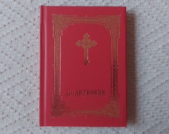 May include: A small red hardcover book featuring a gold embossed cross and an ornate rectangular border on the front cover. The Cyrillic text "МОЛИТВЕННИК" is printed in gold lettering at the bottom center of the cover, indicating it is a prayer book.
