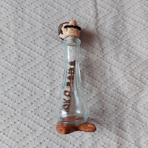 May include: A clear glass bottle with a wooden stopper featuring a cartoon face with a moustache. The bottle has a wooden base and the word "OKOLOBABA" is written on the side.