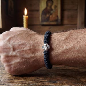 May include: A black knotted cord bracelet featuring a small silver-toned metal cross charm with an embossed religious figure. The bracelet is constructed from thick, textured rope material and is secured around the wrist with a tight, woven knot design.