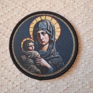 May include: Circular patch depicting a religious image of a woman and child with halos, set against a dark blue backdrop and framed by a black border. The woman wears a blue head covering, and the child is wrapped in a brown cloth.