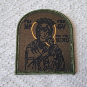 May include: A rectangular patch with a religious icon. The image features a depiction of a woman holding a child, surrounded by a gold halo. The patch has a green border and text in a foreign language.