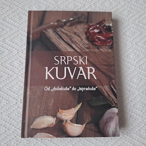 Op de afbeelding: Een bruin kookboek met de titel "SRPSKI KUVAR" in witte tekst. De ondertitel is "Od "dočekuse" do "ispraćuse" in kleinere witte tekst. De omslag toont een houten snijplank met rode pepers, teentjes knoflook en groene kruiden.